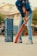 A person wearing a blue sports jersey and jeans is holding an orange cricket bat, appearing ready to play. To the left, there are two stacks of blue Pepsi crates arranged vertically. The background shows a residential building and some greenery.