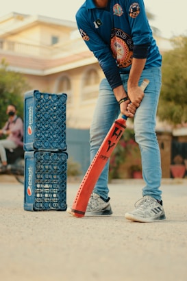 A person wearing a blue sports jersey and jeans is holding an orange cricket bat, appearing ready to play. To the left, there are two stacks of blue Pepsi crates arranged vertically. The background shows a residential building and some greenery.