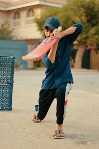 A person wearing a blue shirt and cap, holding a cricket bat, poised to play a shot. They are positioned in an outdoor setting, with a plastic crate nearby, on a paved surface.