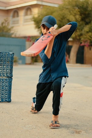 A person wearing a blue shirt and cap, holding a cricket bat, poised to play a shot. They are positioned in an outdoor setting, with a plastic crate nearby, on a paved surface.