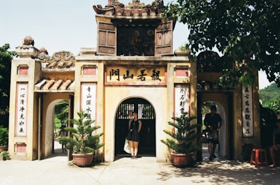 An ornate entrance gate with traditional architectural elements, showing intricate designs and decorative text on pillars. It is surrounded by potted plants, and two people are visible near the entrance. The scene is enriched with lush greenery in the background and a bright, natural setting.