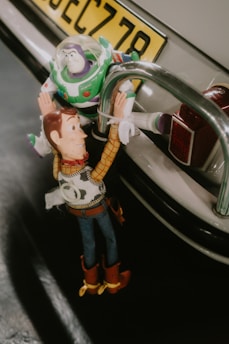 Two toy figures, one dressed as a cowboy and the other as an astronaut, are positioned on the back bumper of a car, with the cowboy hanging onto the astronaut. The scene has a playful and adventurous feel.
