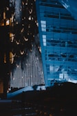 A modern building with a unique, illuminated facade that features vertical light strips. The structure has large glass windows, reflecting the lights and surroundings. There is a darkened area at the bottom with a silhouette of a person standing close to the entrance, which has an illuminated path leading up to it.