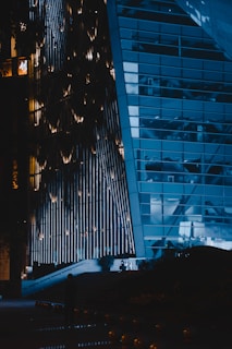 A modern building with a unique, illuminated facade that features vertical light strips. The structure has large glass windows, reflecting the lights and surroundings. There is a darkened area at the bottom with a silhouette of a person standing close to the entrance, which has an illuminated path leading up to it.