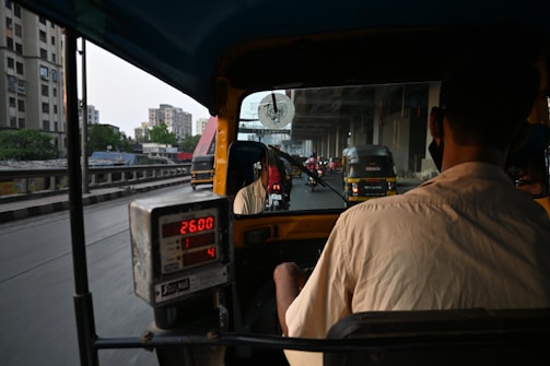 A person is driving an auto rickshaw on a busy urban road. The interior meter reads 26.00, while other vehicles, including more auto rickshaws, are visible on the road. The scene depicts a cityscape with buildings in the background and roads bustling with traffic under an overpass.
