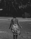 A person wearing a bikini top and a sarong is walking along a sandy beach. The image is in black and white, creating a nostalgic, vintage feel. Palm trees are faintly visible in the background, suggesting a tropical location.