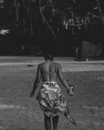 A person wearing a bikini top and a sarong is walking along a sandy beach. The image is in black and white, creating a nostalgic, vintage feel. Palm trees are faintly visible in the background, suggesting a tropical location.