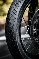 A close-up view of a motorcycle wheel and tire featuring the brand name 'Battlax BT46' etched on the side. The focus is on the tread pattern and the spokes of the wheel, with a blurred greenish background.