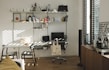 A well-organized home office with a white desk and chair, an ergonomic black chair on wheels, and a computer monitor and laptop. A tote bag hangs from the black chair. The desk is positioned against a wall with multiple wall-mounted shelves filled with books, files, and decorative items. To the right, there's a window with blinds partially open, letting in sunlight, and a wooden cabinet with a coffee machine and a framed poster. The wooden flooring and minimalistic decor create a warm and inviting atmosphere.