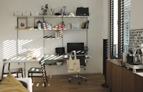 A well-organized home office with a white desk and chair, an ergonomic black chair on wheels, and a computer monitor and laptop. A tote bag hangs from the black chair. The desk is positioned against a wall with multiple wall-mounted shelves filled with books, files, and decorative items. To the right, there's a window with blinds partially open, letting in sunlight, and a wooden cabinet with a coffee machine and a framed poster. The wooden flooring and minimalistic decor create a warm and inviting atmosphere.