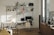 A well-organized home office with a white desk and chair, an ergonomic black chair on wheels, and a computer monitor and laptop. A tote bag hangs from the black chair. The desk is positioned against a wall with multiple wall-mounted shelves filled with books, files, and decorative items. To the right, there's a window with blinds partially open, letting in sunlight, and a wooden cabinet with a coffee machine and a framed poster. The wooden flooring and minimalistic decor create a warm and inviting atmosphere.