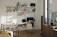 A well-organized home office with a white desk and chair, an ergonomic black chair on wheels, and a computer monitor and laptop. A tote bag hangs from the black chair. The desk is positioned against a wall with multiple wall-mounted shelves filled with books, files, and decorative items. To the right, there's a window with blinds partially open, letting in sunlight, and a wooden cabinet with a coffee machine and a framed poster. The wooden flooring and minimalistic decor create a warm and inviting atmosphere.