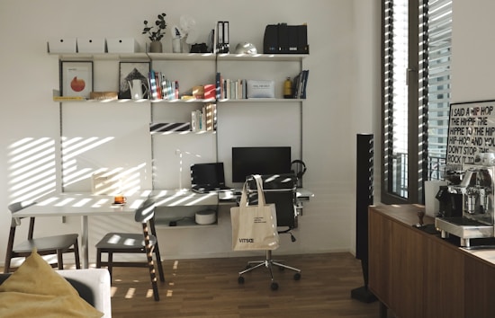 A well-organized home office with a white desk and chair, an ergonomic black chair on wheels, and a computer monitor and laptop. A tote bag hangs from the black chair. The desk is positioned against a wall with multiple wall-mounted shelves filled with books, files, and decorative items. To the right, there's a window with blinds partially open, letting in sunlight, and a wooden cabinet with a coffee machine and a framed poster. The wooden flooring and minimalistic decor create a warm and inviting atmosphere.