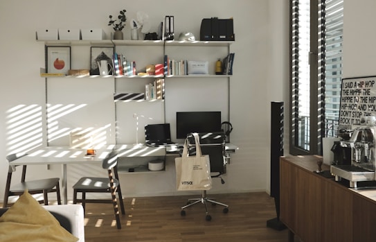 A well-organized home office with a white desk and chair, an ergonomic black chair on wheels, and a computer monitor and laptop. A tote bag hangs from the black chair. The desk is positioned against a wall with multiple wall-mounted shelves filled with books, files, and decorative items. To the right, there's a window with blinds partially open, letting in sunlight, and a wooden cabinet with a coffee machine and a framed poster. The wooden flooring and minimalistic decor create a warm and inviting atmosphere.