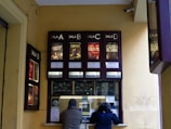 A ticket counter window for a cinema is visible with movie posters displayed above it. There are four sections labeled 'Sala A', 'Sala B', 'Sala C' and 'Sala D'. Two people are interacting with the ticket counter, which has various notices and stickers attached to it.