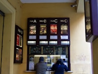 A ticket counter window for a cinema is visible with movie posters displayed above it. There are four sections labeled 'Sala A', 'Sala B', 'Sala C' and 'Sala D'. Two people are interacting with the ticket counter, which has various notices and stickers attached to it.