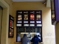 A ticket counter window for a cinema is visible with movie posters displayed above it. There are four sections labeled 'Sala A', 'Sala B', 'Sala C' and 'Sala D'. Two people are interacting with the ticket counter, which has various notices and stickers attached to it.