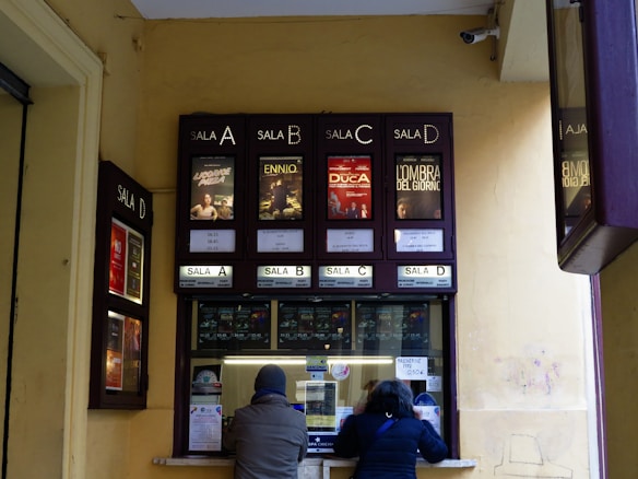 A ticket counter window for a cinema is visible with movie posters displayed above it. There are four sections labeled 'Sala A', 'Sala B', 'Sala C' and 'Sala D'. Two people are interacting with the ticket counter, which has various notices and stickers attached to it.