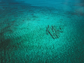 An aerial view of a clear, turquoise body of water with sandy ripples. A shipwreck or structure is visible beneath the surface, casting shadows.