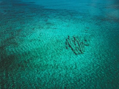 An aerial view of a clear, turquoise body of water with sandy ripples. A shipwreck or structure is visible beneath the surface, casting shadows.