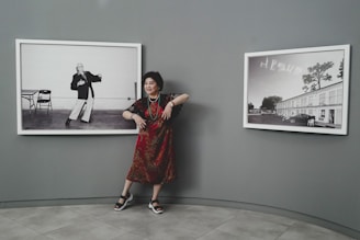 A woman stands in between two black and white framed photographs displayed on a grey wall. She is wearing a red patterned dress, sandals, and some jewelry. The photograph to her left shows a person striking a pose with a table and chair in the background, while the photograph to her right shows a building with the word 'JESUS' written in the sky.