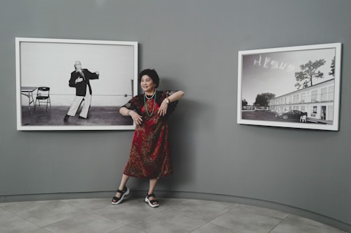 A woman stands in between two black and white framed photographs displayed on a grey wall. She is wearing a red patterned dress, sandals, and some jewelry. The photograph to her left shows a person striking a pose with a table and chair in the background, while the photograph to her right shows a building with the word 'JESUS' written in the sky.