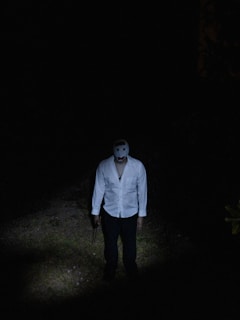 A person standing in a dark environment, wearing a white shirt and a white mask. The figure is illuminated by a spotlight that highlights the ground area and part of the body, creating a suspenseful atmosphere.