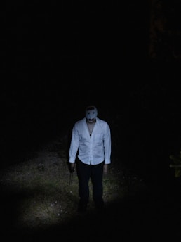 A person standing in a dark environment, wearing a white shirt and a white mask. The figure is illuminated by a spotlight that highlights the ground area and part of the body, creating a suspenseful atmosphere.