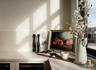 A kitchen countertop with a minimalist decor, featuring a framed still-life painting of oranges, a vase holding branches with white blossoms, black salt and pepper grinders, an open book, and a small white ceramic container. Sunlight casts geometric shadows through a window, enhancing the tranquil atmosphere.