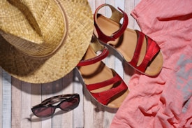 A straw hat, red sandals, a pair of sunglasses, and a pink shirt are arranged on a wooden surface, suggesting a summer vacation theme.