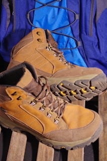 A pair of tan hiking boots with sturdy soles is placed on a wooden surface. Behind the boots, a blue backpack with black straps is visible. The boots have brown laces and metal eyelets.