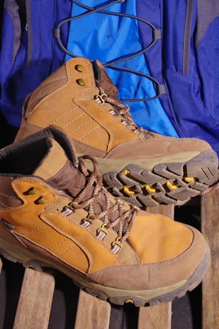 A pair of tan hiking boots with sturdy soles is placed on a wooden surface. Behind the boots, a blue backpack with black straps is visible. The boots have brown laces and metal eyelets.