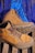 A pair of tan hiking boots with sturdy soles is placed on a wooden surface. Behind the boots, a blue backpack with black straps is visible. The boots have brown laces and metal eyelets.