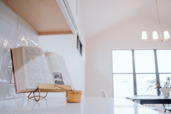 An open cookbook rests on a stand on a white kitchen countertop. Next to it is a brass measuring cup. The background is softly blurred, showing a modern dining area with a chandelier and a window allowing natural light to enter.