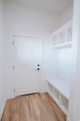 A small, well-lit entryway featuring a closed white door with black hardware on the left side, adjacent to white walls. To the right, there is a built-in white bench with storage cubbies underneath and open shelves above. The flooring is a light wood grain.