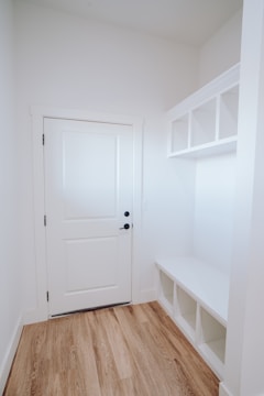 A small, well-lit entryway featuring a closed white door with black hardware on the left side, adjacent to white walls. To the right, there is a built-in white bench with storage cubbies underneath and open shelves above. The flooring is a light wood grain.