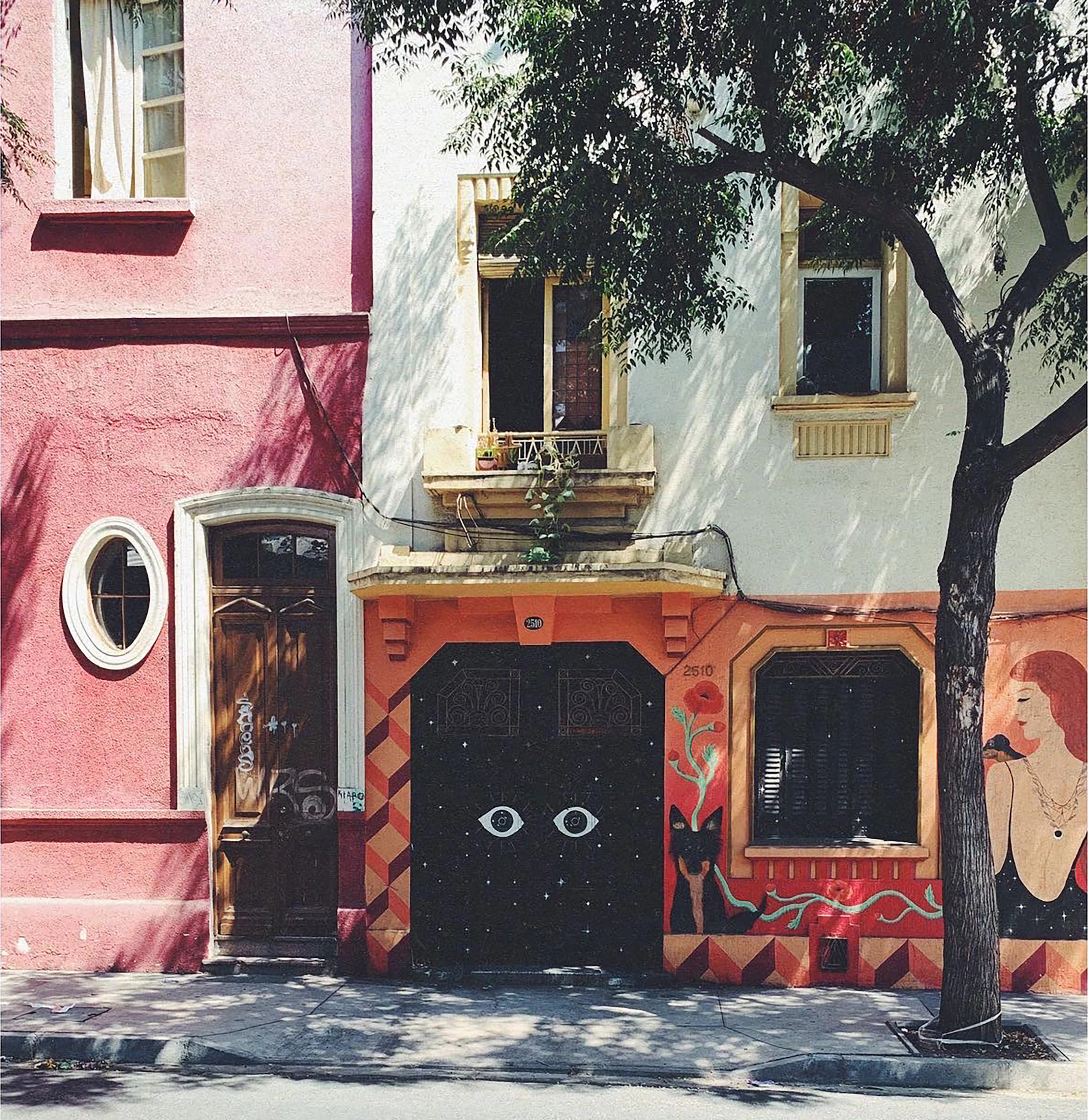 A painted house front in Santiago's Barrio Brasil: a deep pink neighbour with an oval window beside a pale green facade whose black doors carry a mural of two staring eyes and a reclining figure, with a tree throwing shadows across the wall.