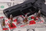 A black handgun is placed on top of a spread of U.S. dollar bills. Surrounding the gun are several bullets standing upright. Visible on the bills are red stains that resemble smeared blood.