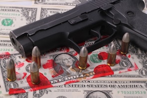 A black handgun is placed on top of a spread of U.S. dollar bills. Surrounding the gun are several bullets standing upright. Visible on the bills are red stains that resemble smeared blood.