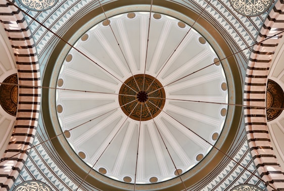 A beautifully intricate dome featuring detailed geometric and floral patterns. The dome's design includes a central medallion with circular motifs and radiating lines. Arabic calligraphy is elegantly incorporated along the perimeter, contributing to the architectural artistry. Neutral tones dominate the design, accentuated by stripes and ornamental elements.