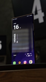 A smartphone displaying a digital clock interface with a dark-themed background. The time reads 16:45, with the word 'Viernes' visible, indicating it's a Friday. The battery status is shown at 60% and there are several app icons at the bottom.