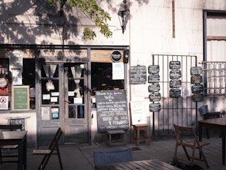 A charming, rustic storefront with a variety of signs and chalkboards displaying menus and notices. The scene includes a weathered wooden door flanked by two small tables with chairs, set in the shade of a tree. There are several blackboards with handwritten menus and advertisements on the wall.