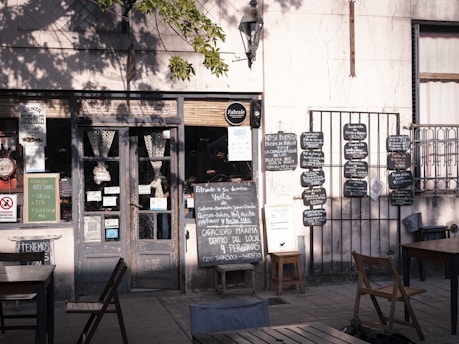 A charming, rustic storefront with a variety of signs and chalkboards displaying menus and notices. The scene includes a weathered wooden door flanked by two small tables with chairs, set in the shade of a tree. There are several blackboards with handwritten menus and advertisements on the wall.