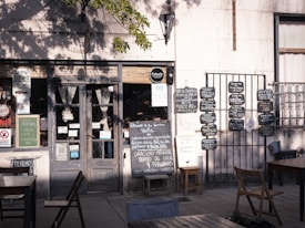 A charming, rustic storefront with a variety of signs and chalkboards displaying menus and notices. The scene includes a weathered wooden door flanked by two small tables with chairs, set in the shade of a tree. There are several blackboards with handwritten menus and advertisements on the wall.