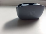 A close-up of a JBL earbud case on a white surface. The case is sleek and dark-colored, with an embossed JBL logo. The lighting casts a soft shadow onto the surface, creating a minimalist aesthetic.