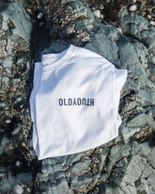 A white T-shirt with black lettering is laid out on a textured rocky surface with barnacles and cracks visible on the stones.