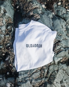 A white T-shirt with black lettering is laid out on a textured rocky surface with barnacles and cracks visible on the stones.
