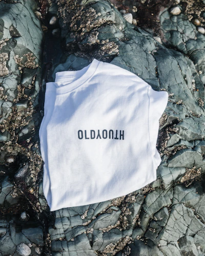 Crisp white premium cotton tee with minimalist black Stark Aura emblem, laid flat on a charcoal surface