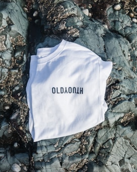 A white T-shirt with black lettering is laid out on a textured rocky surface with barnacles and cracks visible on the stones.