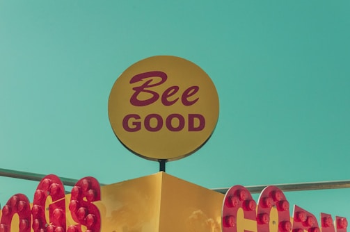 A vintage-style sign features the phrase 'Bee Good' in red letters on a yellow circular background. Red bulbs frame the signage, which sits against a bright turquoise-blue sky.